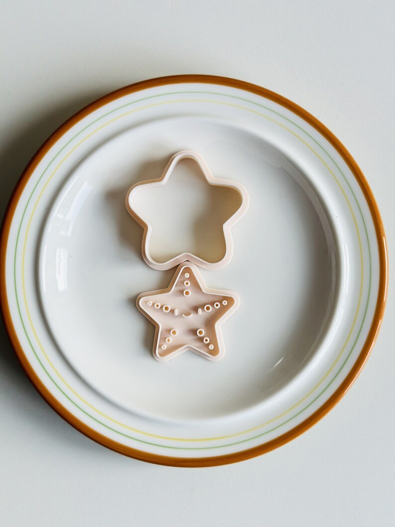 May include: Two plastic cookie cutters in the shape of stars on a white plate with a brown rim. The top star is plain and the bottom star has a face and dots.