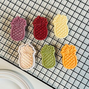 May include: Six pineapple-shaped cookies on a black wire rack. The cookies are in various colors, including red, green, yellow, and white.