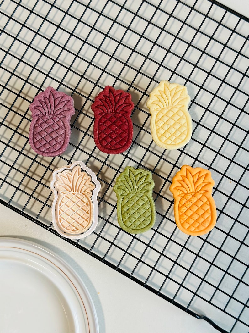 May include: Six pineapple-shaped cookies on a black wire rack. The cookies are in various colors, including red, green, yellow, and white.
