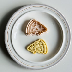 May include: A white ceramic plate with a gold rim, featuring a pink plastic pizza slice cookie cutter and a yellow cookie shaped like a pizza slice.
