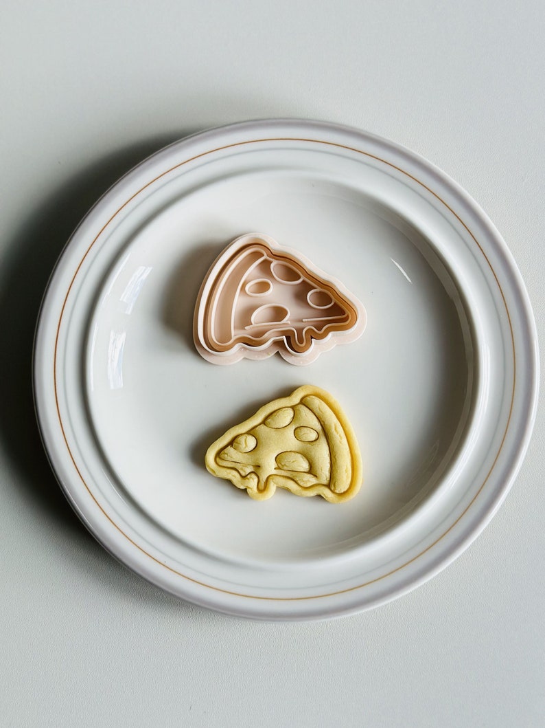 May include: A white ceramic plate with a gold rim, featuring a pink plastic pizza slice cookie cutter and a yellow cookie shaped like a pizza slice.