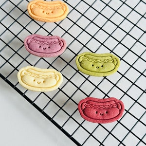 May include: Five hot dog shaped cookies on a black wire rack. The cookies are yellow, purple, green, red, and white. Each cookie has a smiling face.