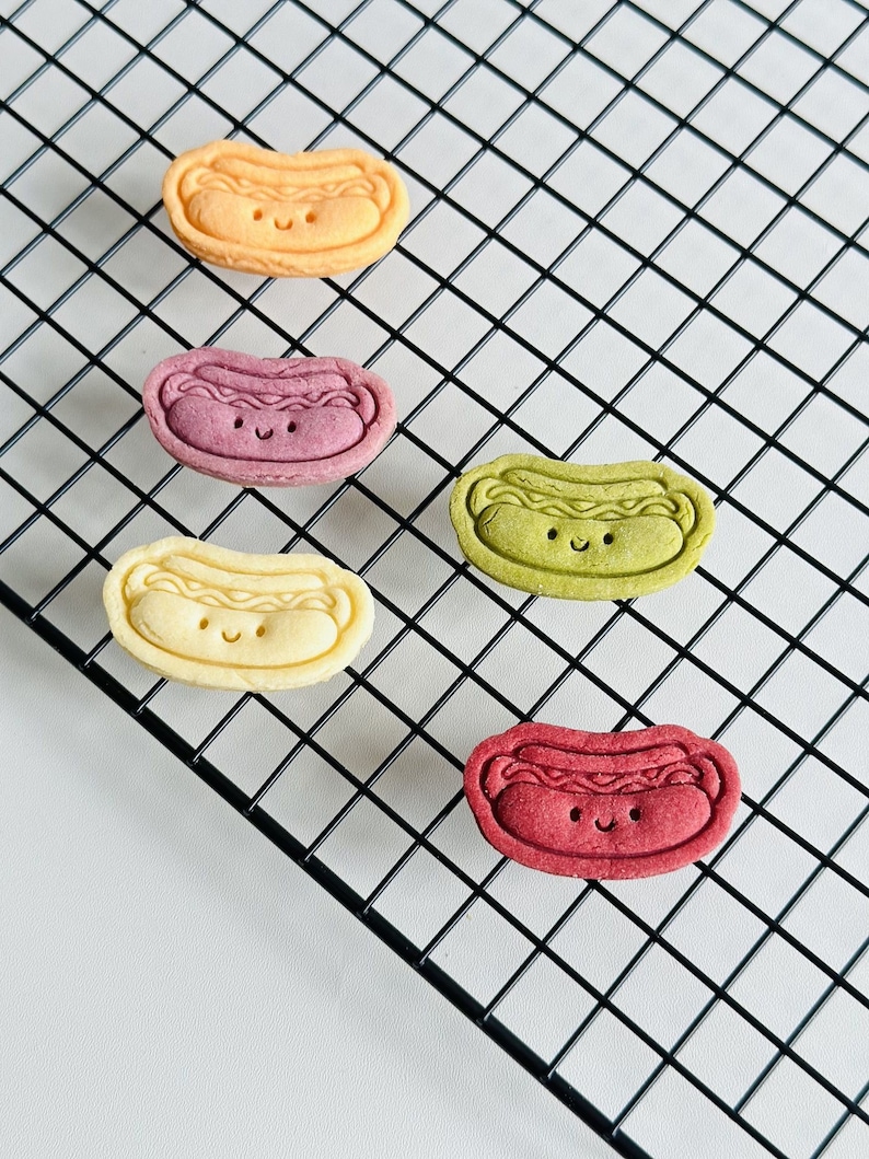 May include: Five hot dog shaped cookies on a black wire rack. The cookies are yellow, purple, green, red, and white. Each cookie has a smiling face.