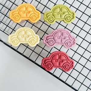 May include: Five colorful car-shaped cookies on a black wire rack. The cookies are yellow, green, white, purple, and red.