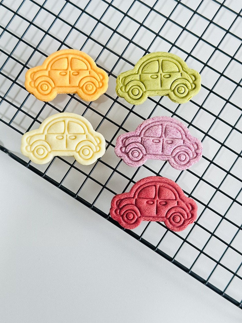 May include: Five colorful car-shaped cookies on a black wire rack. The cookies are yellow, green, white, purple, and red.