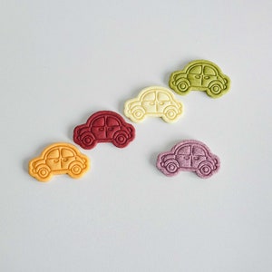 May include: Five small car-shaped cookies in different colors: yellow, red, white, green, and purple. The cookies are arranged in a line on a light gray background.
