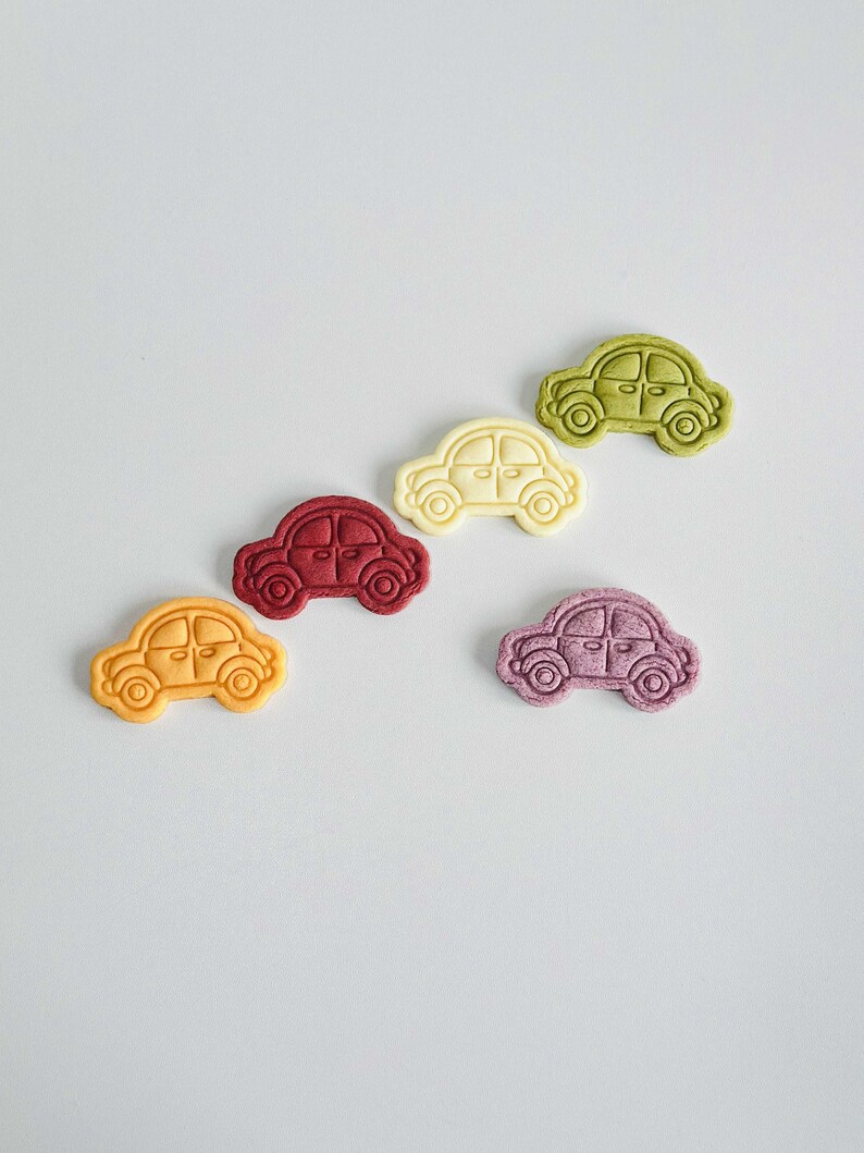 May include: Five small car-shaped cookies in different colors: yellow, red, white, green, and purple. The cookies are arranged in a line on a light gray background.