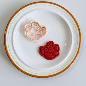 Op de afbeelding: Een witte plaat met een bruine en gele rand bevat een roze kuikenvormige koekjesvorm en een rood kuikenvormig koekje. De koekjesvorm is van plastic en het koekje is van rood deeg.