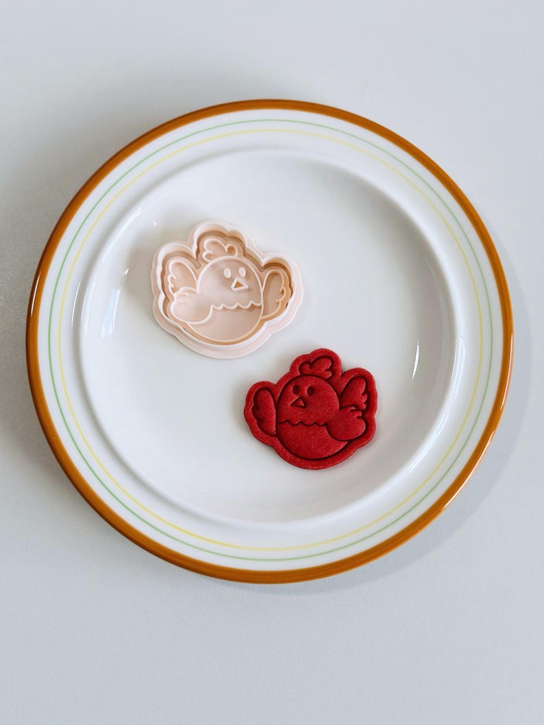 Op de afbeelding: Een witte plaat met een bruine en gele rand bevat een roze kuikenvormige koekjesvorm en een rood kuikenvormig koekje. De koekjesvorm is van plastic en het koekje is van rood deeg.