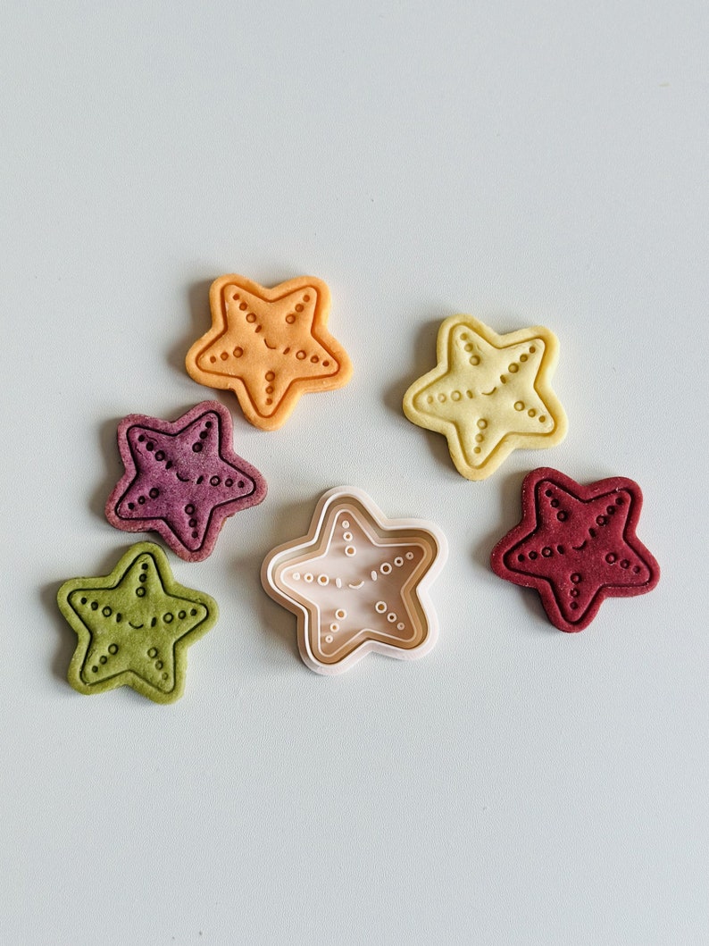 May include: Five colourful star-shaped cookies with a smiling face and a white plastic cookie cutter in the shape of a star with a smiling face.