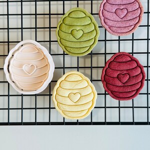 Pu&ograve; includere: Cinque biscotti a forma di ape, uno in uno stampo per biscotti, su una griglia di filo metallico nera. I biscotti sono di vari colori: verde, rosa, rosso e giallo. Ogni biscotto ha un piccolo cuore al centro.