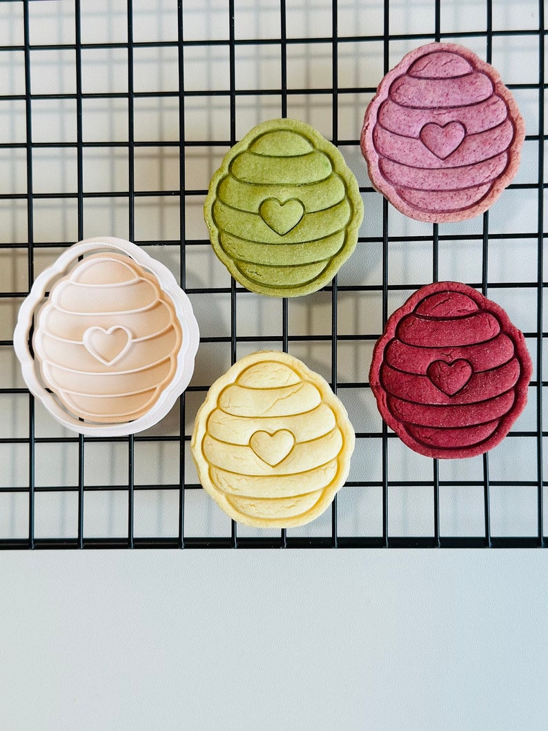 Pu&ograve; includere: Cinque biscotti a forma di ape, uno in uno stampo per biscotti, su una griglia di filo metallico nera. I biscotti sono di vari colori: verde, rosa, rosso e giallo. Ogni biscotto ha un piccolo cuore al centro.