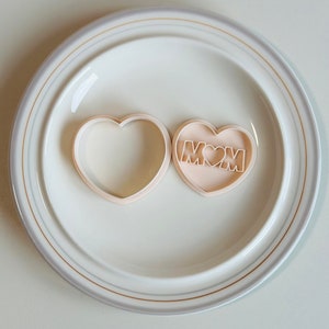 May include: Two heart-shaped cookie cutters, one plain and one with the word "MOM" and a small heart inside. The cutters are on a white plate with a gold rim.