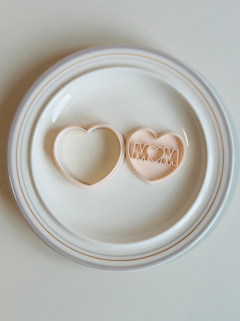 May include: Two heart-shaped cookie cutters, one plain and one with the word "MOM" and a small heart inside. The cutters are on a white plate with a gold rim.