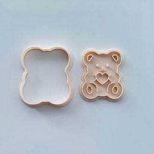 May include: A beige plastic cookie cutter in the shape of a bear holding a heart. The cutter is shown next to its matching outline cutter.