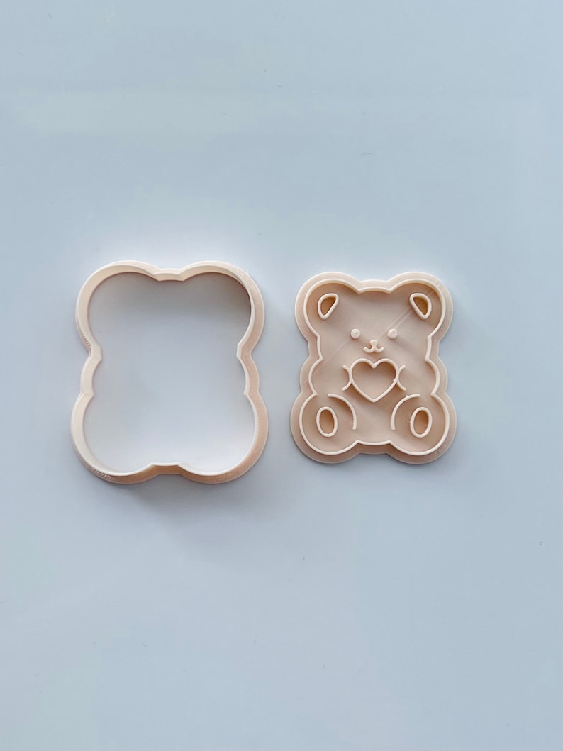 May include: A beige plastic cookie cutter in the shape of a bear holding a heart. The cutter is shown next to its matching outline cutter.
