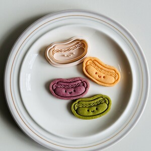 May include: Four hot dog shaped cookies on a white plate. The cookies are different colors: pink, orange, purple, and green. Each cookie has a smiling face.