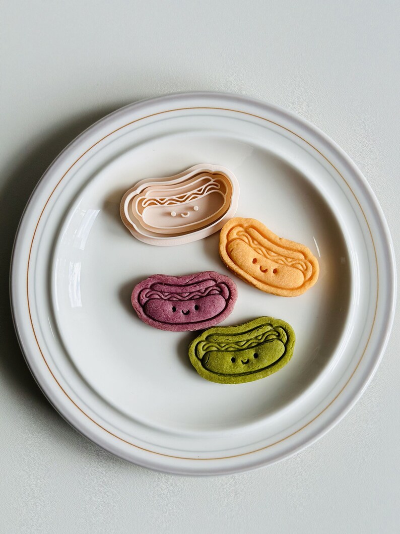 May include: Four hot dog shaped cookies on a white plate. The cookies are different colors: pink, orange, purple, and green. Each cookie has a smiling face.