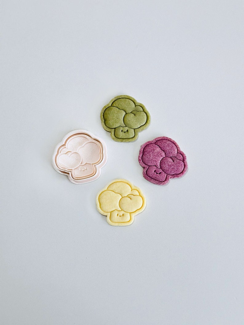 May include: Four broccoli-shaped cookies, one green, one pink, one yellow, and one purple, with a smiling face on each.