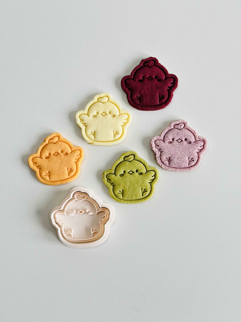 Op de afbeelding: Zes koekjes in de vorm van kuikens in verschillende kleuren, waaronder oranje, geel, groen, roze en bordeaux. Elk koekje heeft een hartdetail en ligt op een witte achtergrond. De koekjes zien er vers gebakken uit.