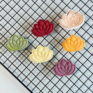 May include: Six lotus flower-shaped cookies in various colors, including red, green, yellow, and purple, on a black wire cooling rack.