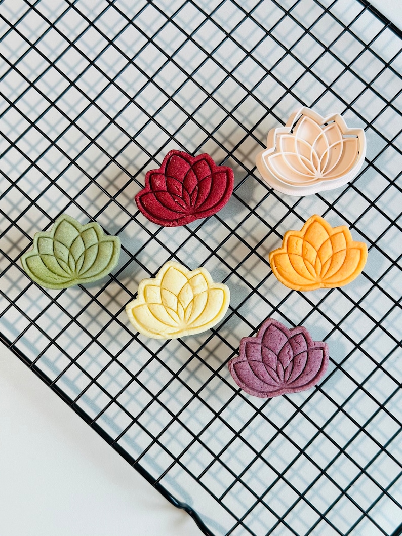 May include: Six lotus flower-shaped cookies in various colors, including red, green, yellow, and purple, on a black wire cooling rack.