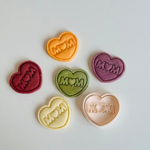 May include: Five heart-shaped cookies with the word "MOM" and a heart in the center. The cookies are in various colors, including red, green, yellow, and white. There is also a white heart-shaped cookie cutter with the same design.