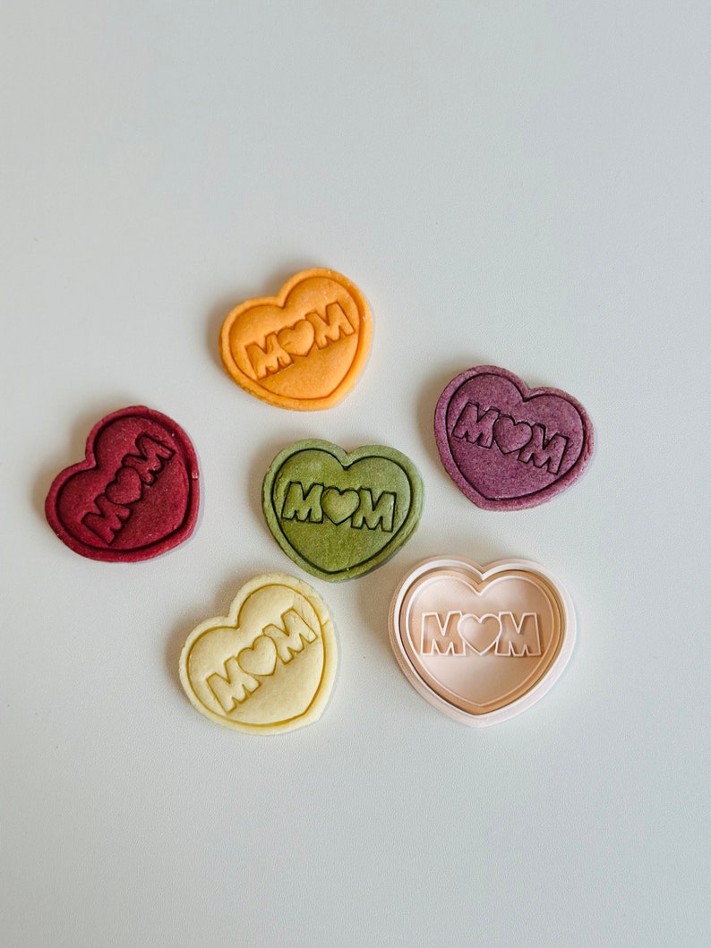 May include: Five heart-shaped cookies with the word "MOM" and a heart in the center. The cookies are in various colors, including red, green, yellow, and white. There is also a white heart-shaped cookie cutter with the same design.