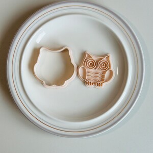 May include: Two plastic owl-shaped cookie cutters on a white plate with a gold rim. One cutter is a simple outline of an owl, while the other has detailed features, including eyes, beak, and feathers.