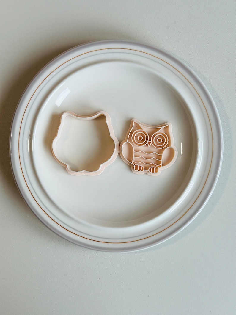 May include: Two plastic owl-shaped cookie cutters on a white plate with a gold rim. One cutter is a simple outline of an owl, while the other has detailed features, including eyes, beak, and feathers.