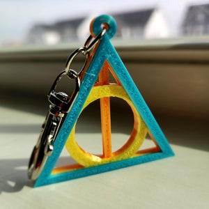 May include: A 3D-printed keychain featuring a blue triangle with an orange and yellow circle and bar design. The keychain has a silver clasp and chain. The design is a symbol from a popular fantasy series.