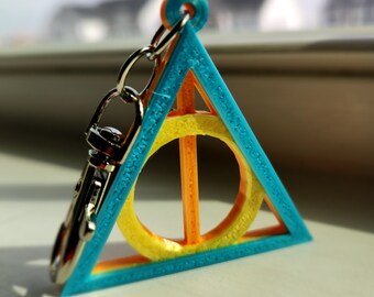 May include: A 3D-printed keychain featuring a blue triangle with an orange and yellow circle and bar design. The keychain has a silver clasp and chain. The design is a symbol from a popular fantasy series.