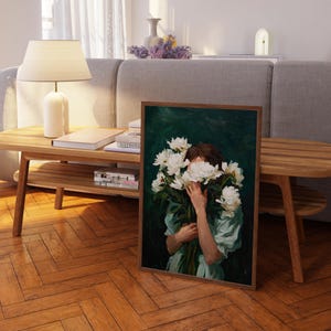 a painting of a woman holding a bouquet of white flowers, with her face partially obscured by the flowers. The painting is displayed on a wooden table in a living room setting, with a lamp and other decorative items visible in the background.