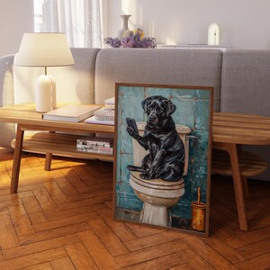 a painting of a black dog sitting on a toilet, holding a cell phone in its mouth. The painting is displayed on a wooden table in a living room setting.