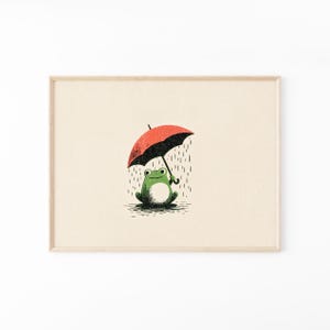 以下が含まれることがあります： 赤い傘と黒い傘をさして雨の中に座る緑色のカエルのイラストが描かれた額入りプリント。明るいベージュの背景の中央に配置されており、手描き風の質感と、降り注ぐ雨を表す垂直の線が特徴的なデザインとなっています。