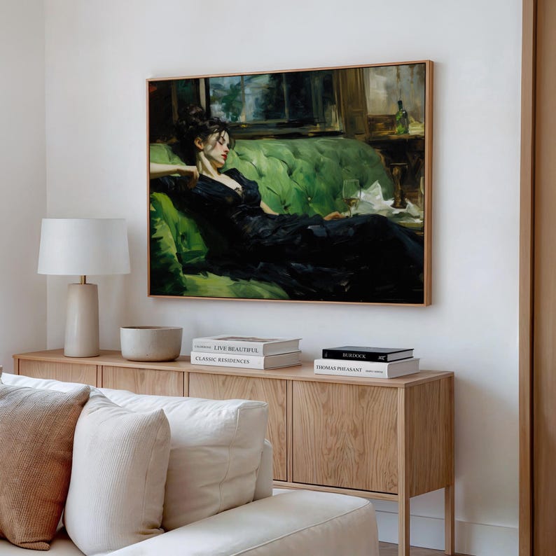a large painting of a woman in a black dress sitting on a green couch, with a lamp and books on a wooden side table below the painting.