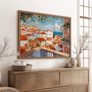 a large framed painting of a colorful cityscape with buildings, balconies, and trees, hanging on a wall above a wooden dresser.