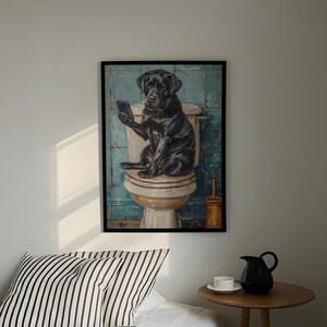 A framed painting of a black dog sitting on a toilet, holding a phone, is hanging on a wall above a bed with a striped pillow.