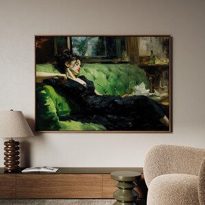 a woman in a black dress sitting on a green couch, with a lamp and a book on a side table in front of her.