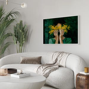 a modern living room with a white sofa, a wooden side table, and a framed painting of a woman with yellow flowers covering her face.