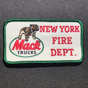 Könnte beinhalten: Ein weißer gestickter Aufnäher mit grünem Rand. Der Aufnäher zeigt einen roten Bulldoggen, das Mack Trucks Logo und den Text "NEW YORK FIRE DEPT."