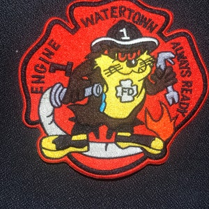 Op de afbeelding: Rode, zwarte en gele geborduurde patch met een cartoonkat die een brandweerhelm draagt. De patch heeft de tekst "Watertown Engine 1 Always Ready" en heeft een brandkraan op de borst van de kat.