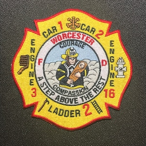 May include: A yellow and red fire department patch with the words "Worcester", "Engine 3", "Ladder 2", "Car 1", "Car 2", "Engine 16", "A Step Above The Rest", "Courage", and "Compassion". The patch features a firefighter holding a baby in his arms.