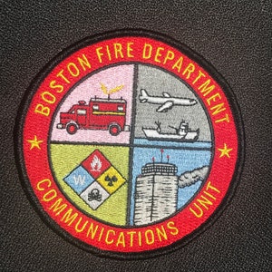 May include: A red, yellow, and black embroidered patch with the words "Boston Fire Department Communications Unit" around the edge. The patch is divided into four quadrants, each with a different symbol: a fire truck, an airplane, a building, and a symbol with a skull and crossbones.