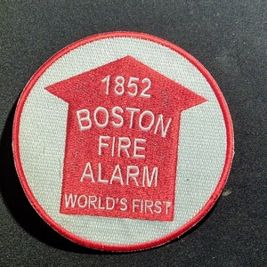 Può includere: Un cerotto circolare rosso e bianco con il testo "1852 Boston Fire Alarm World's First" in lettere bianche.