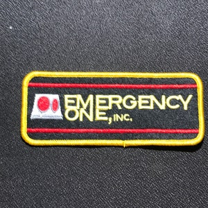 Könnte beinhalten: Ein gestickter Aufnäher in Gelb und Rot mit dem Text "EMERGENCY ONE, INC." und einem roten und weißen Logo eines stilisierten Gebäudes.