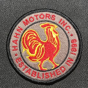 Könnte beinhalten: Ein gestickter grauer und schwarzer Aufnäher mit einem roten Hahn und dem Text "Hahn Motors Inc. Gegründet im Jahr 1889."