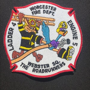 以下が含まれることがあります： 赤い縁取りのある白い刺繍パッチ。パッチには、ラダーを持ったコヨーテとホースを持ったロードランナーの漫画が描かれています。「LADDER 4」、「WORCESTER FIRE DEPT.」、「ENGINE 5」、「WEBSTER SQ.」、「THE ROADRUNNERS」の文字がパッチに刺繍されています。