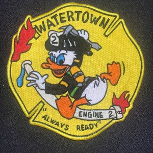 May include: Yellow and red embroidered patch featuring a cartoon duck wearing a firefighter's hat and holding a hose. The patch reads "Watertown Engine 2 Always Ready".