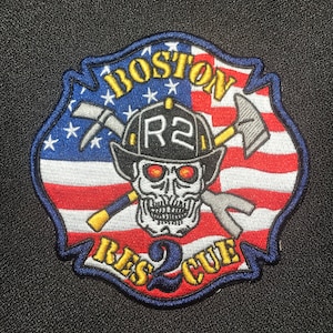 May include: A red, white and blue American flag patch with a skull and crossbones in the centre. The skull is wearing a black firefighter helmet with the letters "R2" on the front. The patch is surrounded by a blue border and the words "BOSTON RESCUE" in yellow letters.